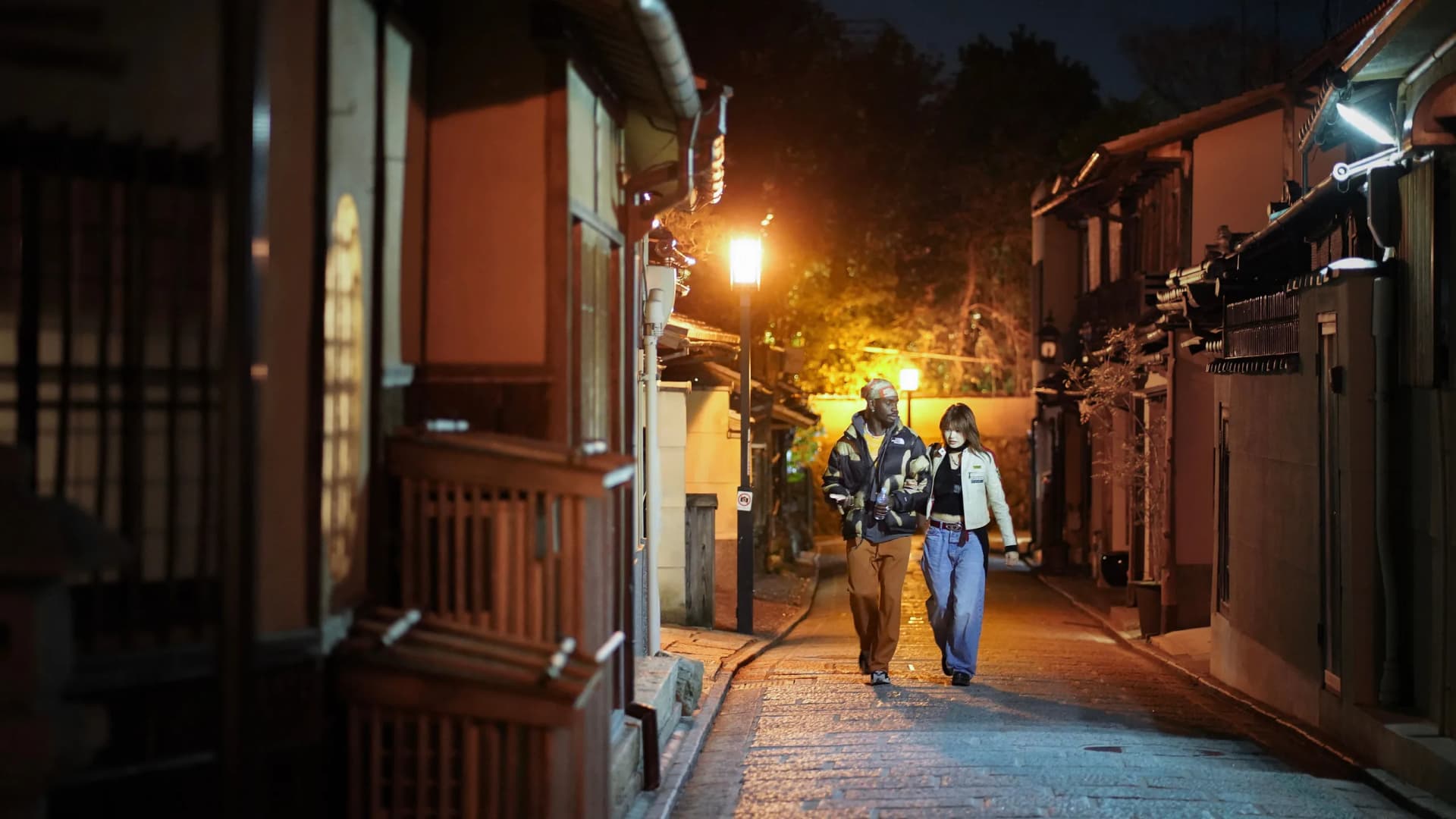Two travelers walking along a lantern-lit street lined with traditional buildings in Japan at night